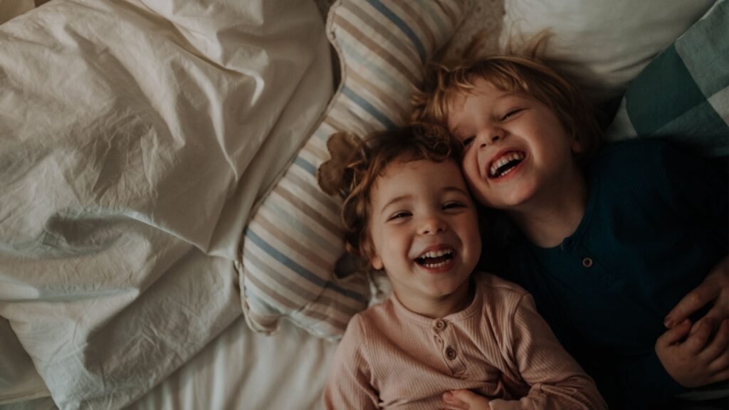 Candid portrait of siblings lying together in bed, looking at the camera and laughing. Top view.