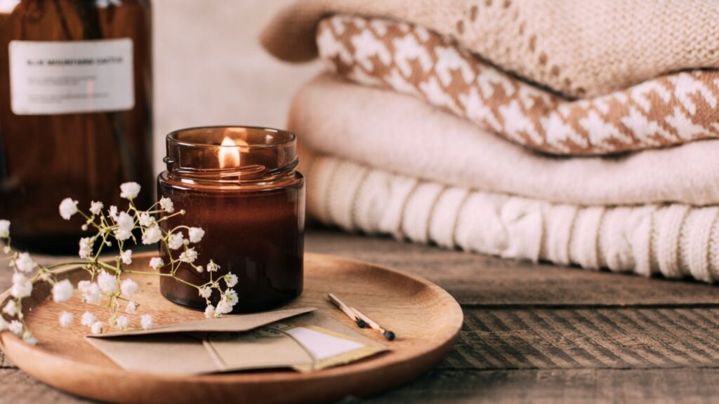 Burning candle in small amber glass jar, flowers of gypsophila and stack knitted seasin sweaters. Cozy lifestyle, hygge concept