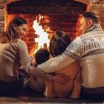 Happy young family parents with two small kids gathering around brick fireplace in cozy and warm country house