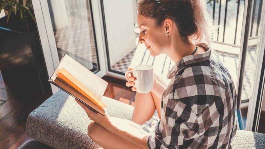 Young pretty woman sitting at opened window drinking coffee and reading a book enjoys of rest