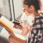 Young pretty woman sitting at opened window drinking coffee and reading a book enjoys of rest