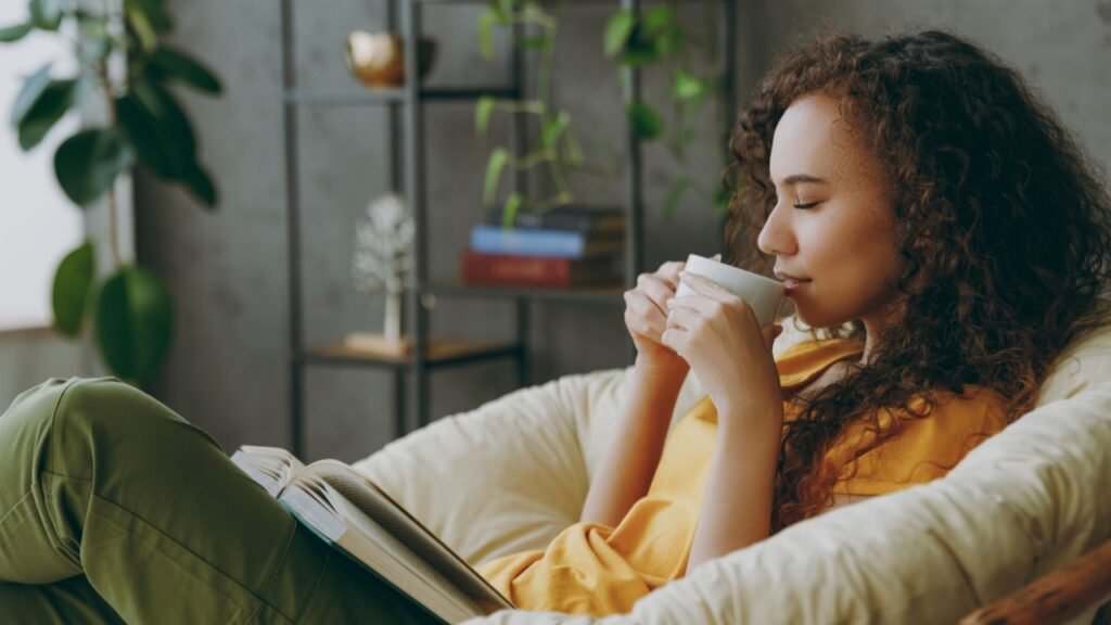 Sideways young woman of African American ethnicity wear t-shirt drink coffee read book close eyes sits in armchair stay at home flat rest relax spend free spare time in living room indoor grey wall