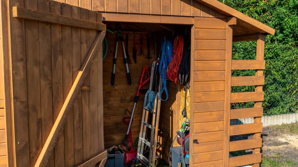 A wooden garden shed standing on a concrete foundation in a garden, visible garden tools.