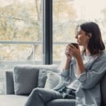 Cheerful dreamy girl holding mug of hot beverage, looking away in deep good thoughts, smiling, dreaming, enjoying aromatic herbal tea, smelling morning coffee, warming hands