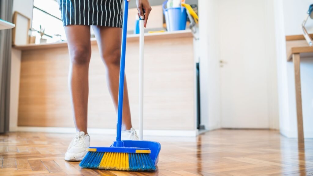 Portrait of young afro woman sweeping wooden floor with broom at home. Cleaning, housework and housekeeping concept.