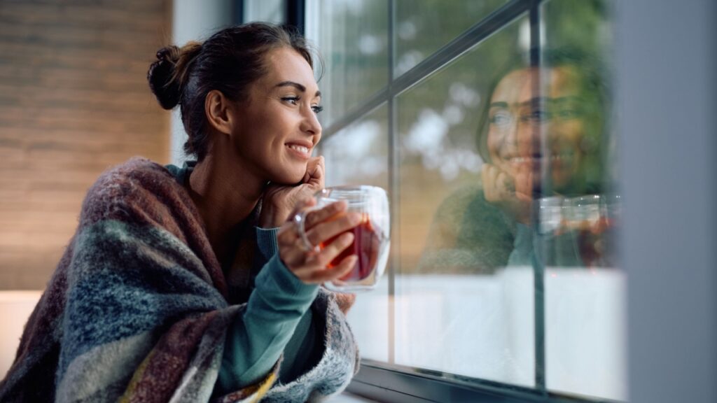 Young smiling woman drinking tea and looking through the window at home. Copy space.