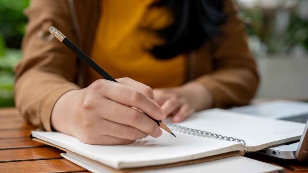 A close-up image of an Asian woman holding a pencil, writing something in her notebook, making list, drawing, taking notes, doing homework, or keeping diary.