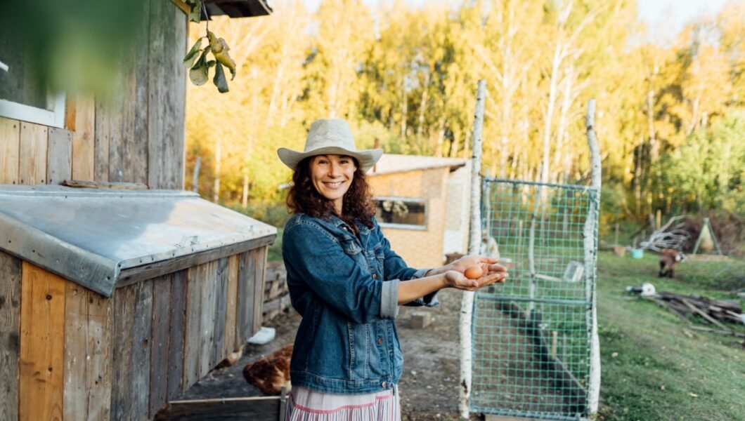 A curly-haired woman in farm clothes beams as she takes an egg from a chicken coop. A woman shows true happiness with a fresh egg in her hand while standing on her farm