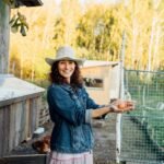 A curly-haired woman in farm clothes beams as she takes an egg from a chicken coop. A woman shows true happiness with a fresh egg in her hand while standing on her farm