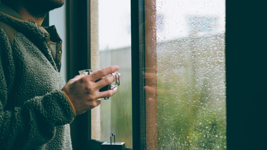 A guy at home holding a cup of coffee looking outside the window on a rainy day