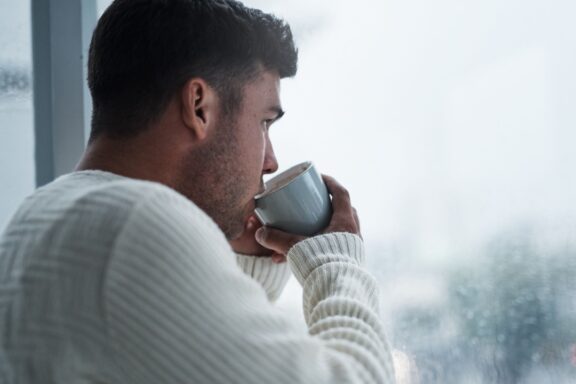 A simple break will bring you back to you. Shot of a young man having coffee and looking out of a window on a rainy day at home.