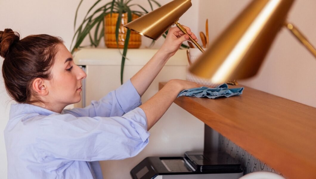 A young woman in a blue shirt is dusting off a wooden shelf in the kitchen. A girl in casual clothes wipes a lamp in the dining room at home. The teenager cleans the house. Modern and cozy interior