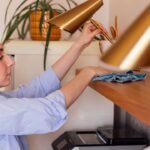 A young woman in a blue shirt is dusting off a wooden shelf in the kitchen. A girl in casual clothes wipes a lamp in the dining room at home. The teenager cleans the house. Modern and cozy interior