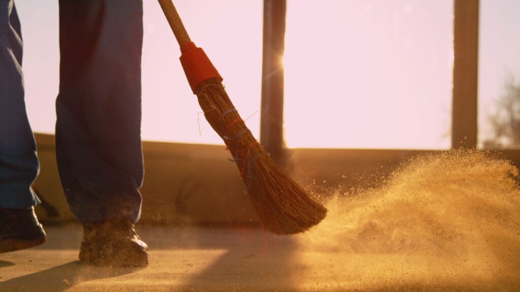 CLOSE UP, LENS FLARE, LOW ANGLE_ Contractor is sweeping the dirty floor with a retro straw broom at scenic golden sunset. Worker sweeps the dusty floor after a long day at a busy construction site