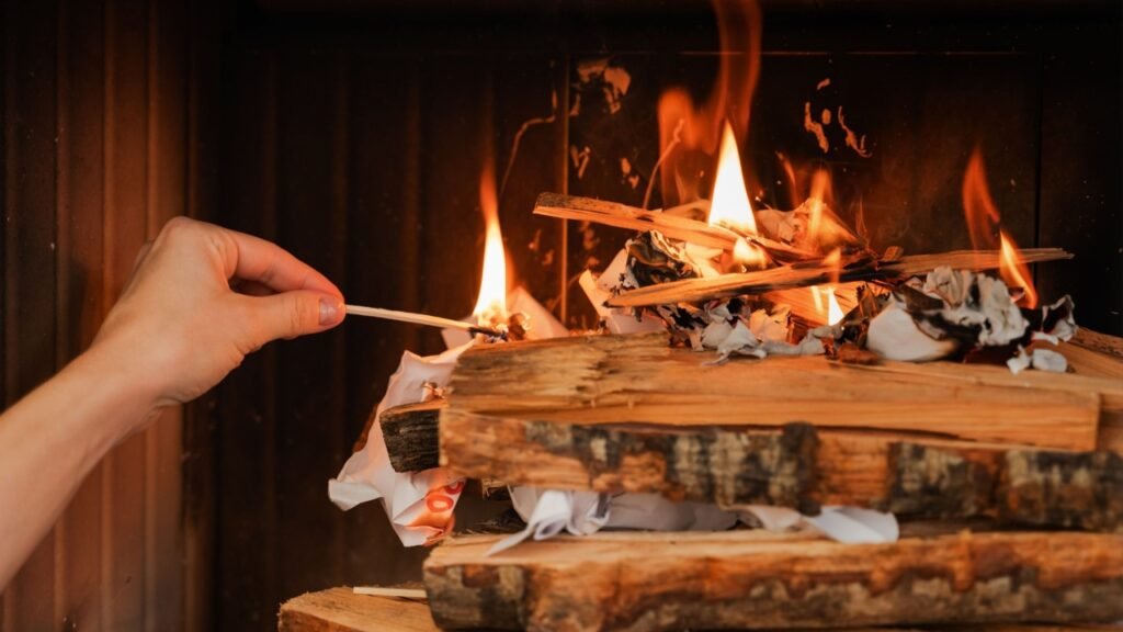 Female hand holds a matchstick, ready to ignite the firewood in a fireplace.