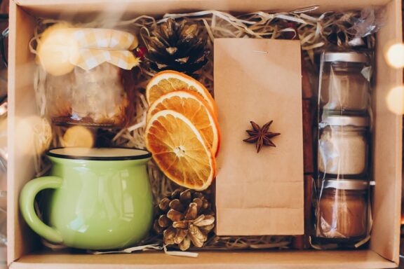 Gift box with tea set. Tea, cup, jam or honey, spices, dried oranges, anise in box on wooden table with christmas lights, top view. Beautiful present, care package for family and friends