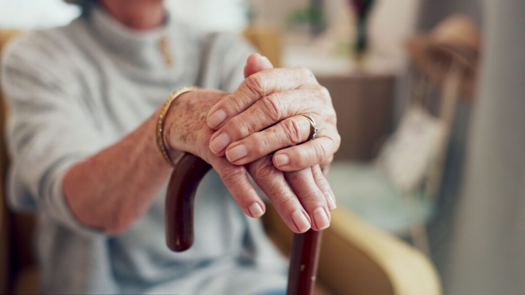 Hands, walking stick and sofa in home for senior person with disability, mobility and recovery from stroke. Elderly care, cane and support for steps, balance and relax on living room couch in house