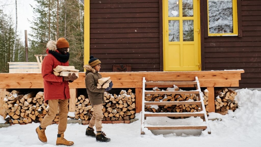 Happy young black man and little boy in warm casual winterwear carrying firewood to their country house located in the forest