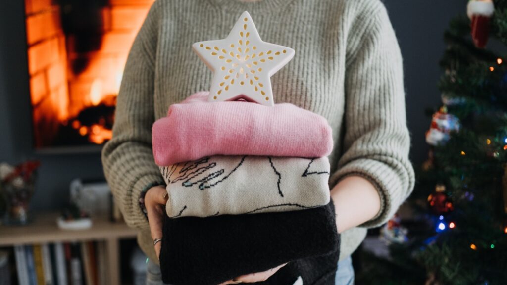 Person holding a stack of folded clothes with ceramic star decoration, cozy Christmas setting. Decluttering during holiday season with fireplace in the background.