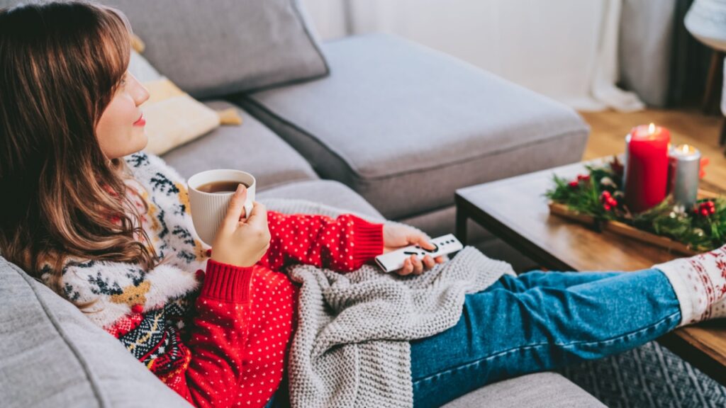 Relaxed woman in plaid and warm socks drinking hot tea with remote controller on sofa at home while watching movie, TV with christmas decoration atmosphere. Cozy and comfortable winter concept