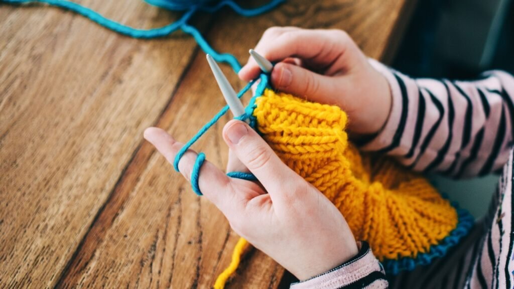 Young girl knitting a circle scarf with yellow and blue coloured yarn. Sitting at the wooden table, close up of the knitting needles.